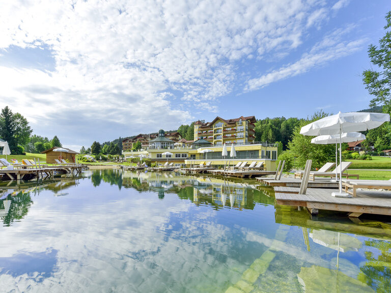 Der Blick auf das Hotel Mooshof über den glänzenden Naturbadesee.